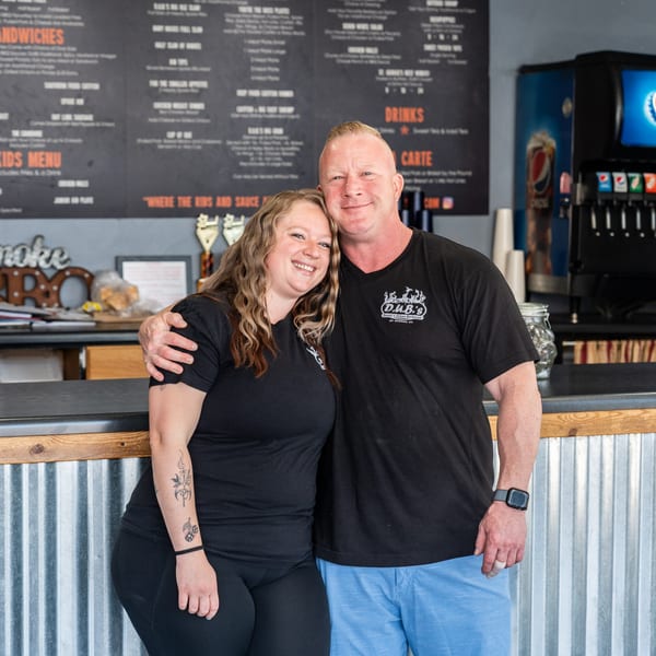 Smiling couple in black D.U.B.'s Urban Barbecue shirts standing in front of a menu board.