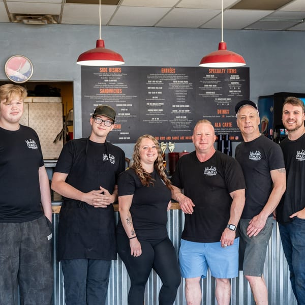 Six smiling D.U.B.'s BBQ employees, including men and a woman, stand in front of a menu board.
