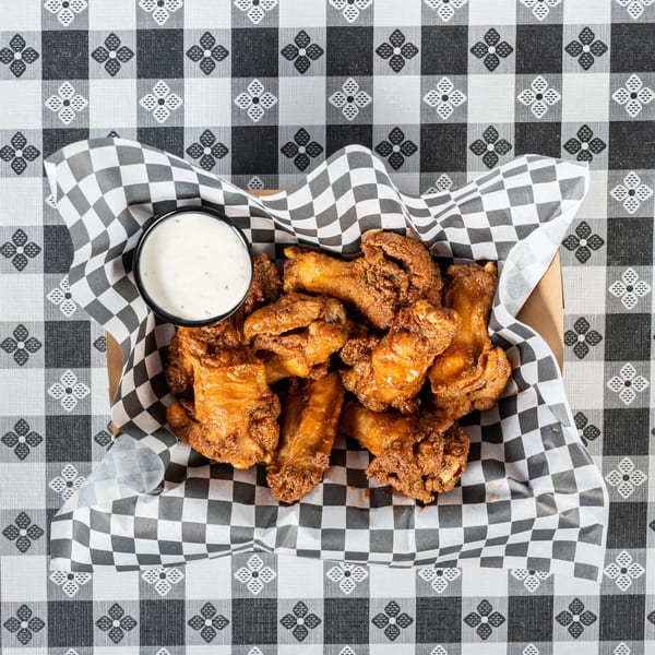 Crispy fried chicken wings with a side of ranch dressing in a basket on a checkered tablecloth.